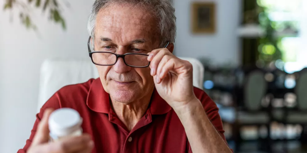 Elderly man in a red shirt focuses on reading a medicine bottle, adjusting glasses. He appears thoughtful in a cozy, sunlit living room.