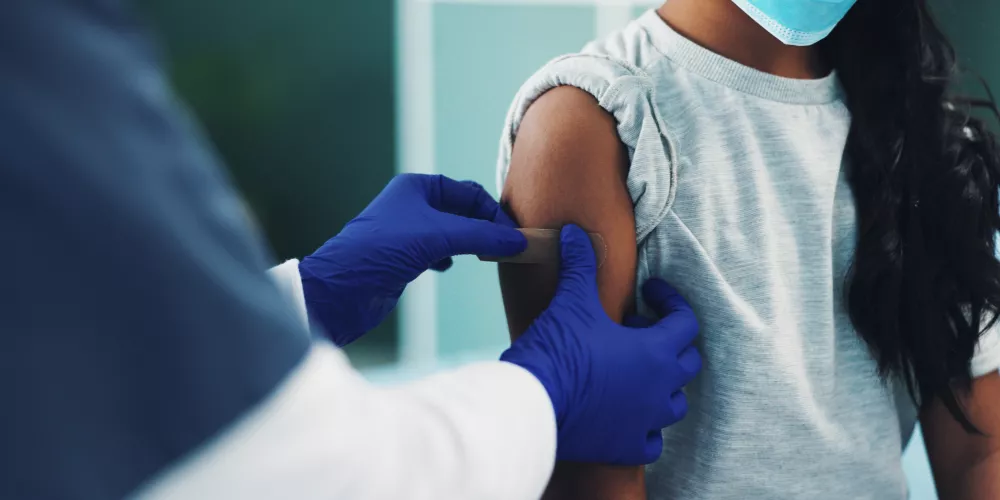 A healthcare worker with blue gloves places a bandage on a child's arm after a vaccination. The child wears a face mask and a gray shirt, conveying a sense of care and safety.