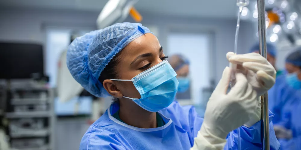 Female anesthesiologist adjusts IV drip in sterile operating room. She is concentrated and careful. Medical staff and equipment appear in the blurred background.