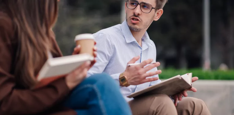 Two young professionals are sitting on a bench, discussing work and reviewing notes in their notebooks